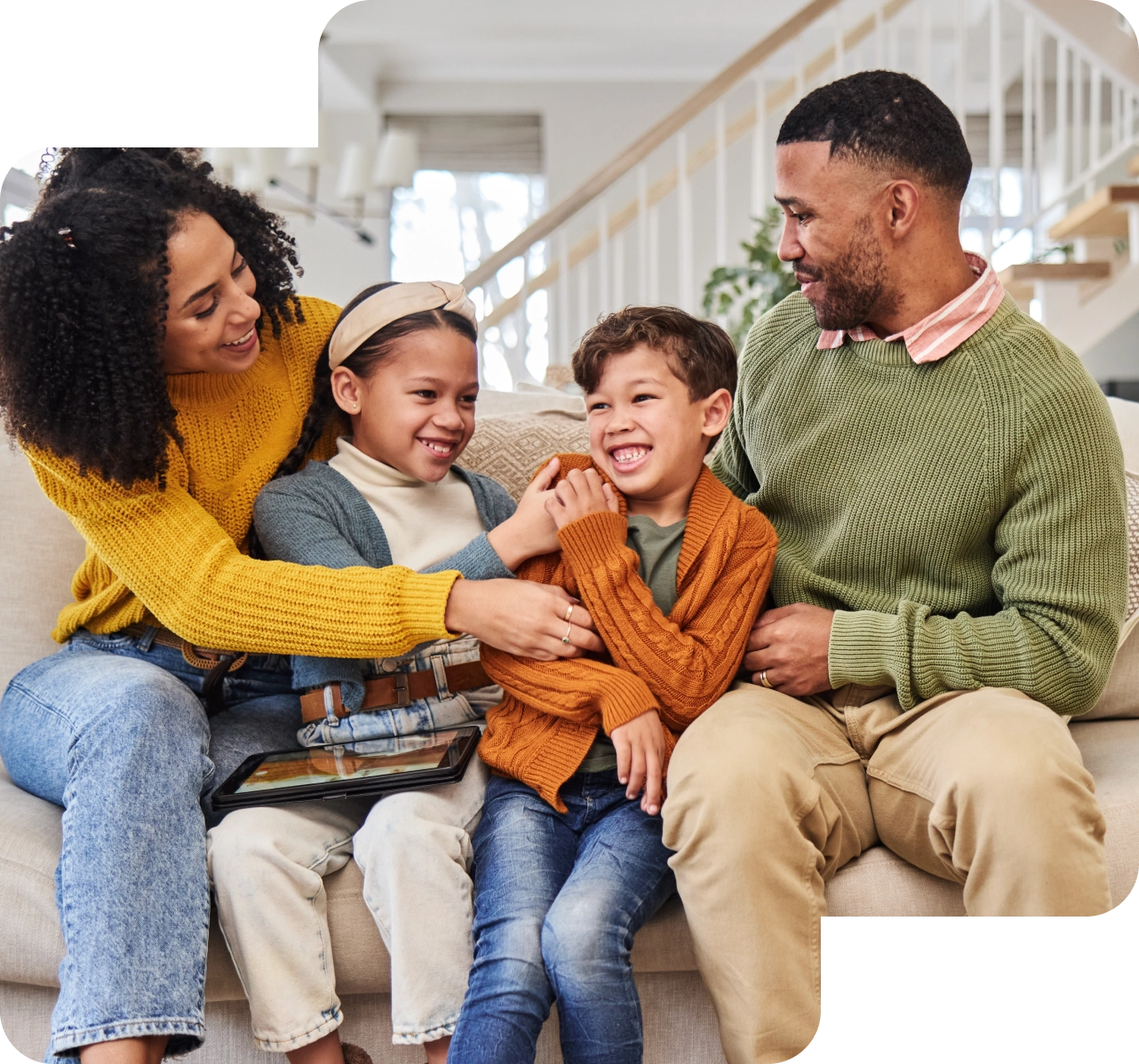 A family of four sits on a couch in a beautifully staged living room, smiling and interacting joyfully. The adult woman wears a yellow sweater, and the man sports a green one. Their children snuggle between them, perfectly embodying the essence of a dream home in this real estate gem.