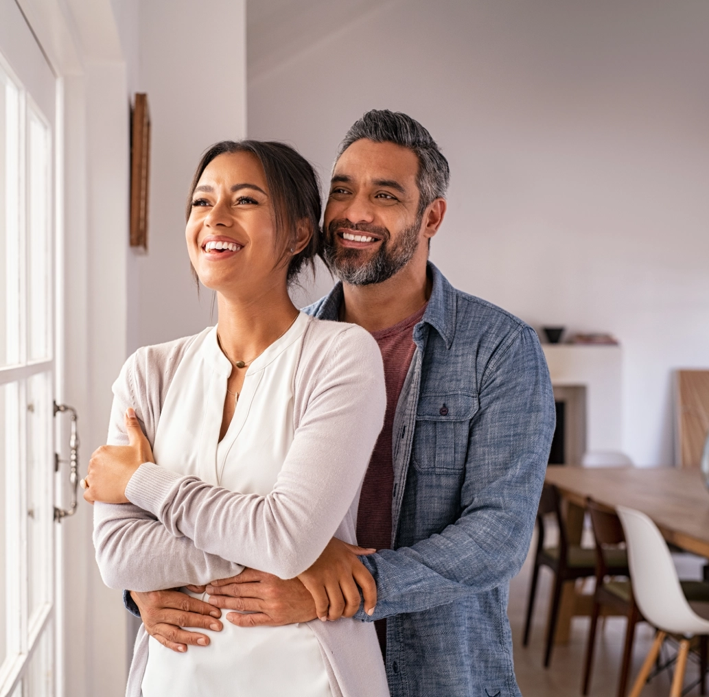 A couple stands smiling in a bright room, embracing the excitement of buying a house. The woman is in front with the man's arms around her as they stand near a large window, basking in the sunlight and dreaming of their future together.