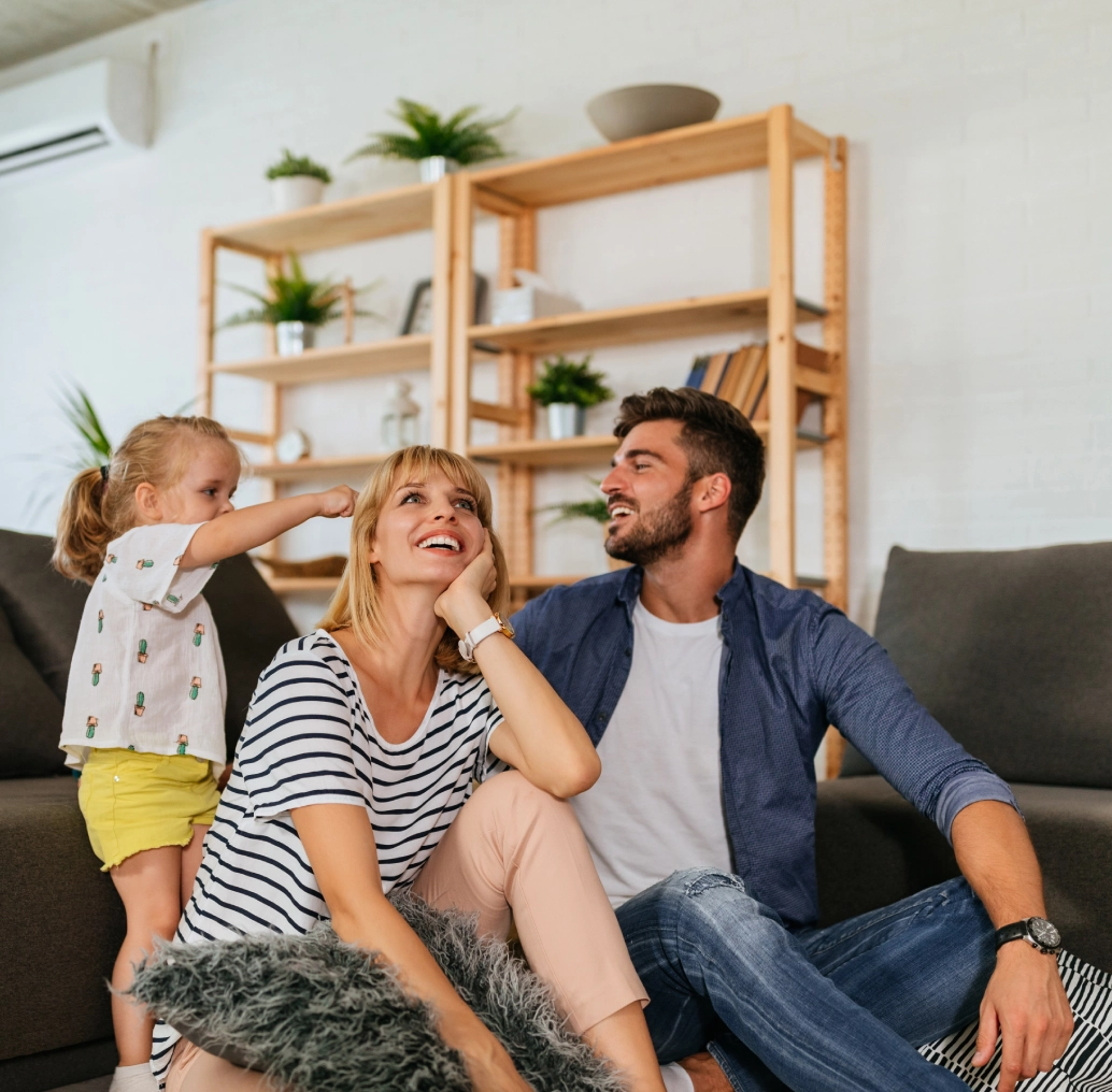 A family of three sits on a couch in their cozy new living room. The child stands joyfully on the couch, while the woman and man sit on the floor, smiling at each other, cherishing the excitement of buying a house together.